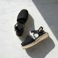 A pair of black woven espadrille wedge sandals with ankle straps and gold-tone buckles, arranged on a white marble surface with strong diagonal shadows.