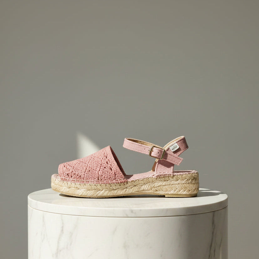 A single pink woven espadrille sandal with an ankle strap and braided jute platform sole, displayed on a white marble cylinder pedestal against a neutral gray background.