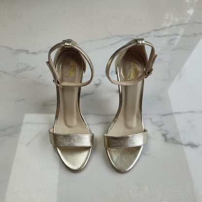 Pair of gold high-heeled sandals on a marble surface with green leaves in the background