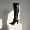 A single black leather knee-high boot with a block heel and pointed toe, photographed against a white studio backdrop with a warm beige wall visible behind it.