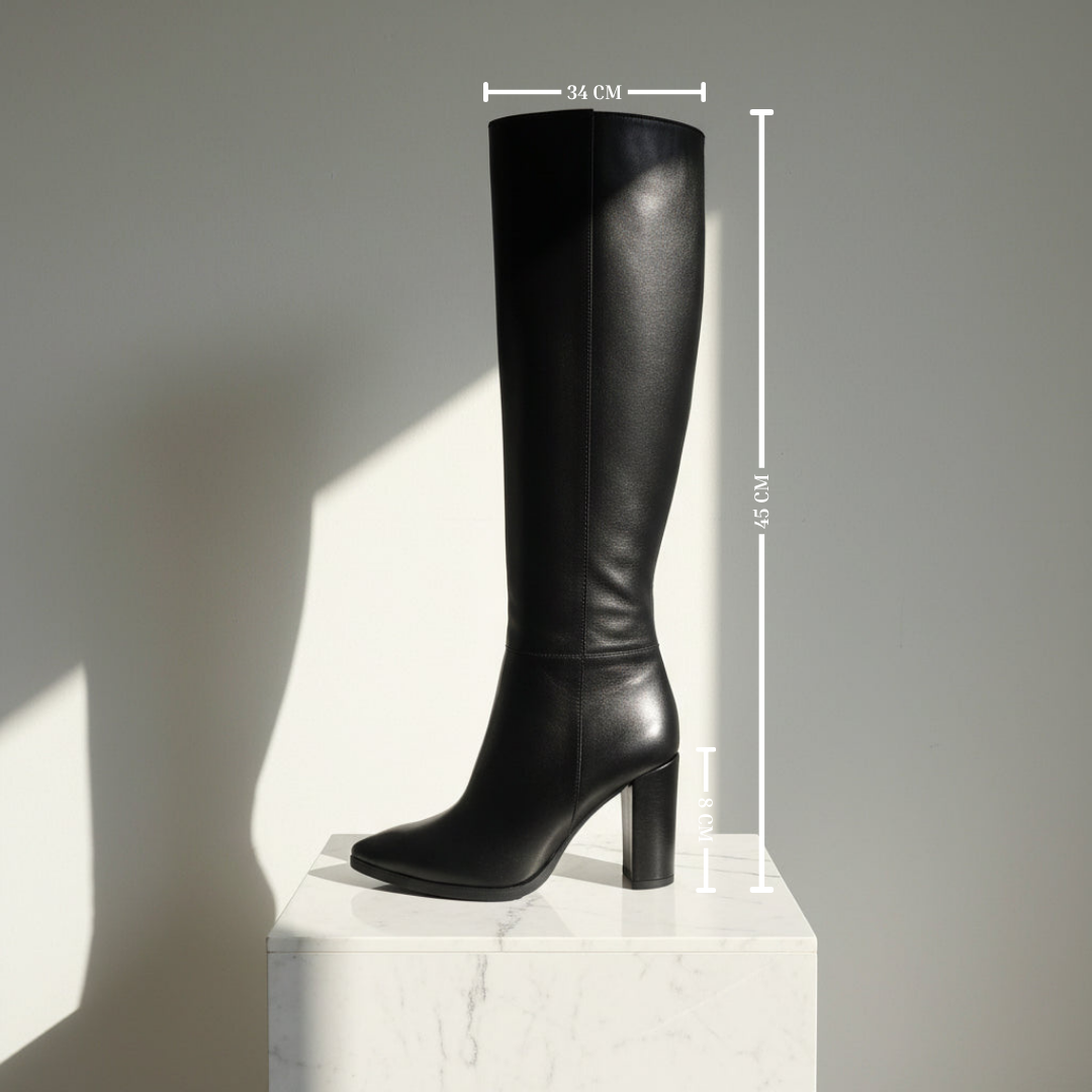 A single black leather knee-high boot with a block heel and pointed toe, shown on a white marble pedestal against a gray wall with dimension lines indicating height and width measurements.