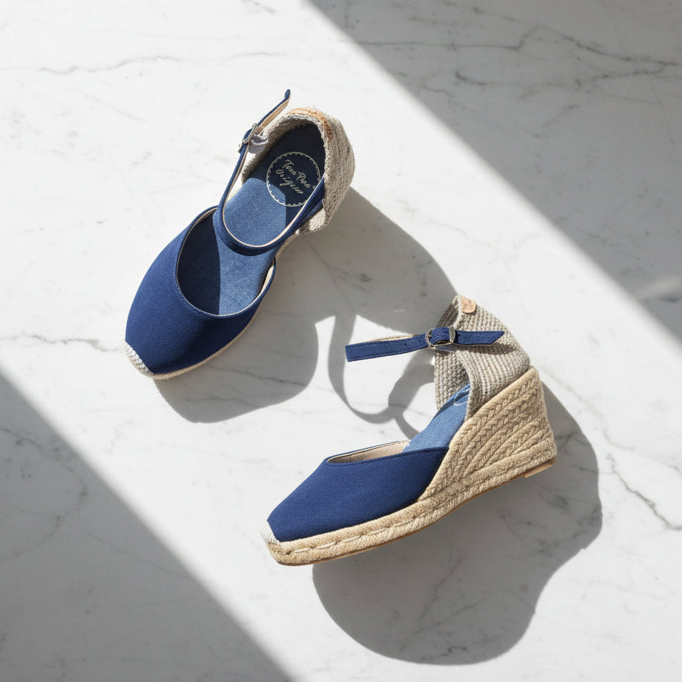 A pair of blue wedge espadrille sandals with woven jute heels and ankle straps, displayed on a white marble surface with diagonal shadows.