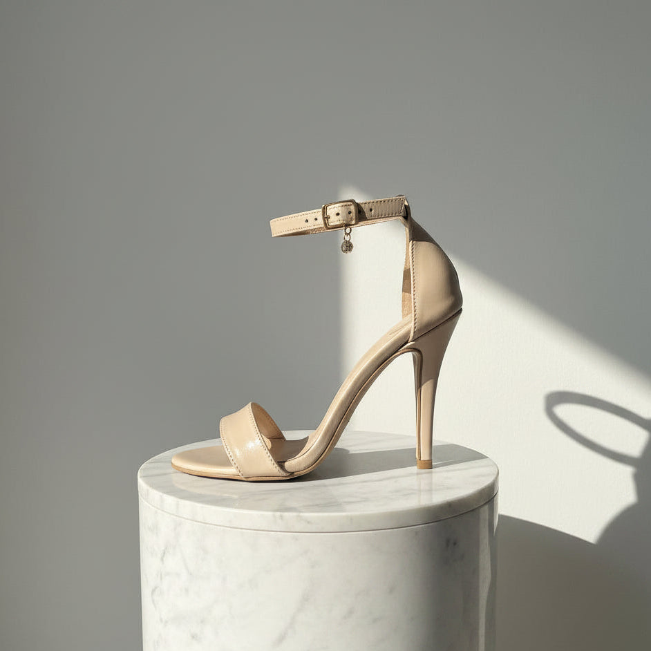 A single beige high-heeled sandal with an ankle strap and small dangling charm, photographed on a light surface against a neutral indoor background.