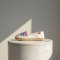 A single slip-on espadrille shoe with a white canvas upper, colorful striped fabric accents in pink, blue, and red, and a natural jute-wrapped sole, displayed on a white marble cylinder pedestal against a gray background.