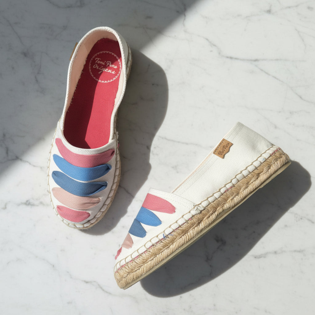 A pair of espadrille slip-on shoes with a white canvas upper, colorful wavy stripe pattern in pink, blue, and beige, and a natural jute-wrapped sole, displayed on a white marble surface.