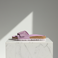 A single lavender-purple slide sandal with a cork midsole and white outsole, displayed on a white marble pedestal against a gray background with a diagonal light shadow.
