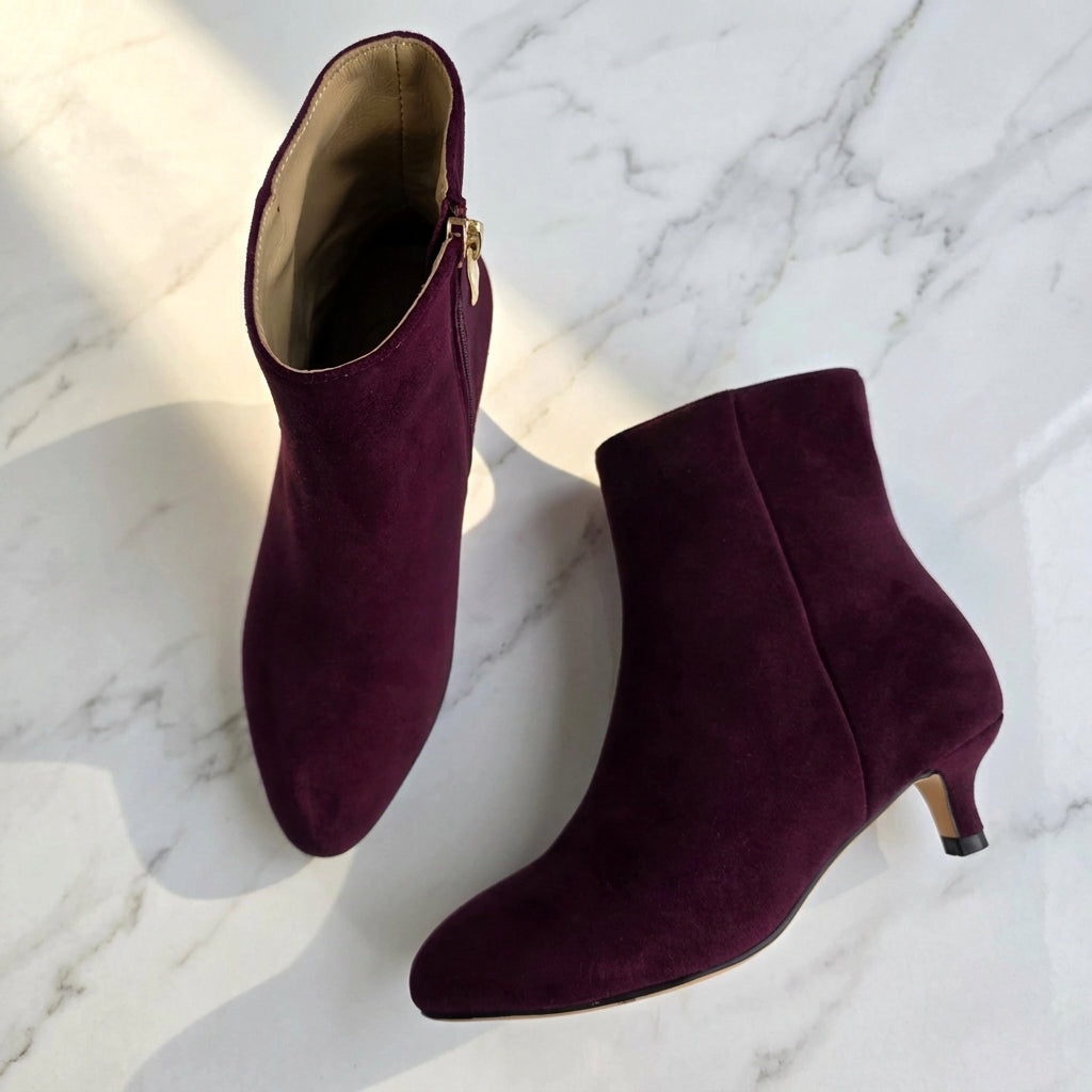 A pair of women's pointed-toe ankle boots in deep burgundy suede, displayed on a white surface. One boot is shown from the side, highlighting the gold zipper, and the other is shown from the top, displaying the low kitten heel.