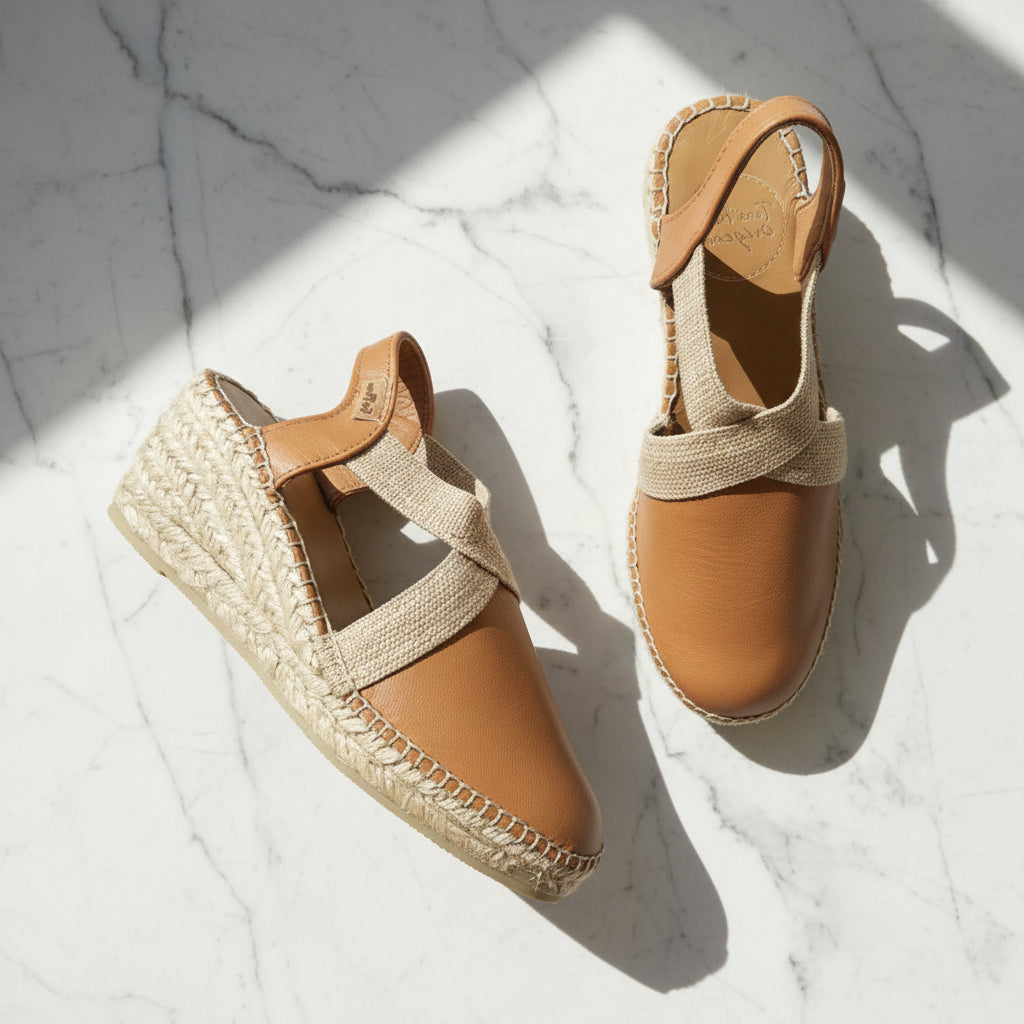 A pair of women's espadrille wedge sandals with tan leather uppers and woven jute soles, arranged on a white marble surface with diagonal shadows.