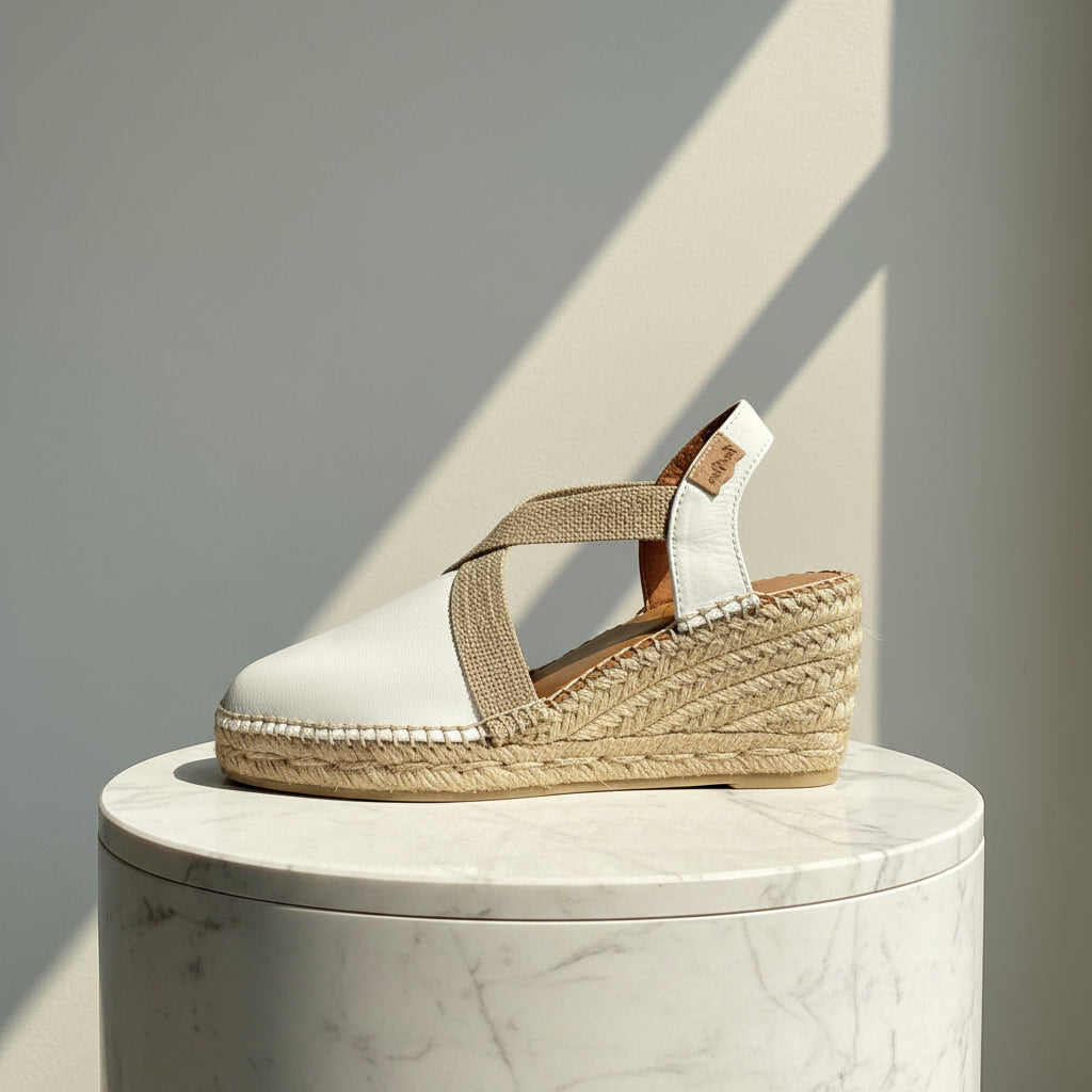 A single white espadrille wedge sandal with woven jute straps and a white ankle strap, displayed on a round white marble pedestal against a gray wall with diagonal shadows.