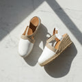 A pair of white espadrille wedge sandals with beige fabric straps, displayed on a light gray marble surface under diagonal shadows.