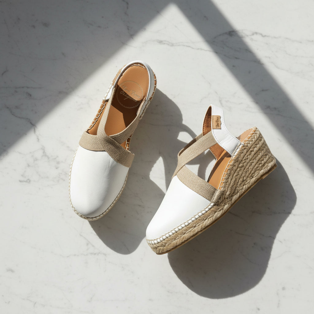 A pair of white espadrille wedge sandals with beige fabric straps, displayed on a light gray marble surface under diagonal shadows.