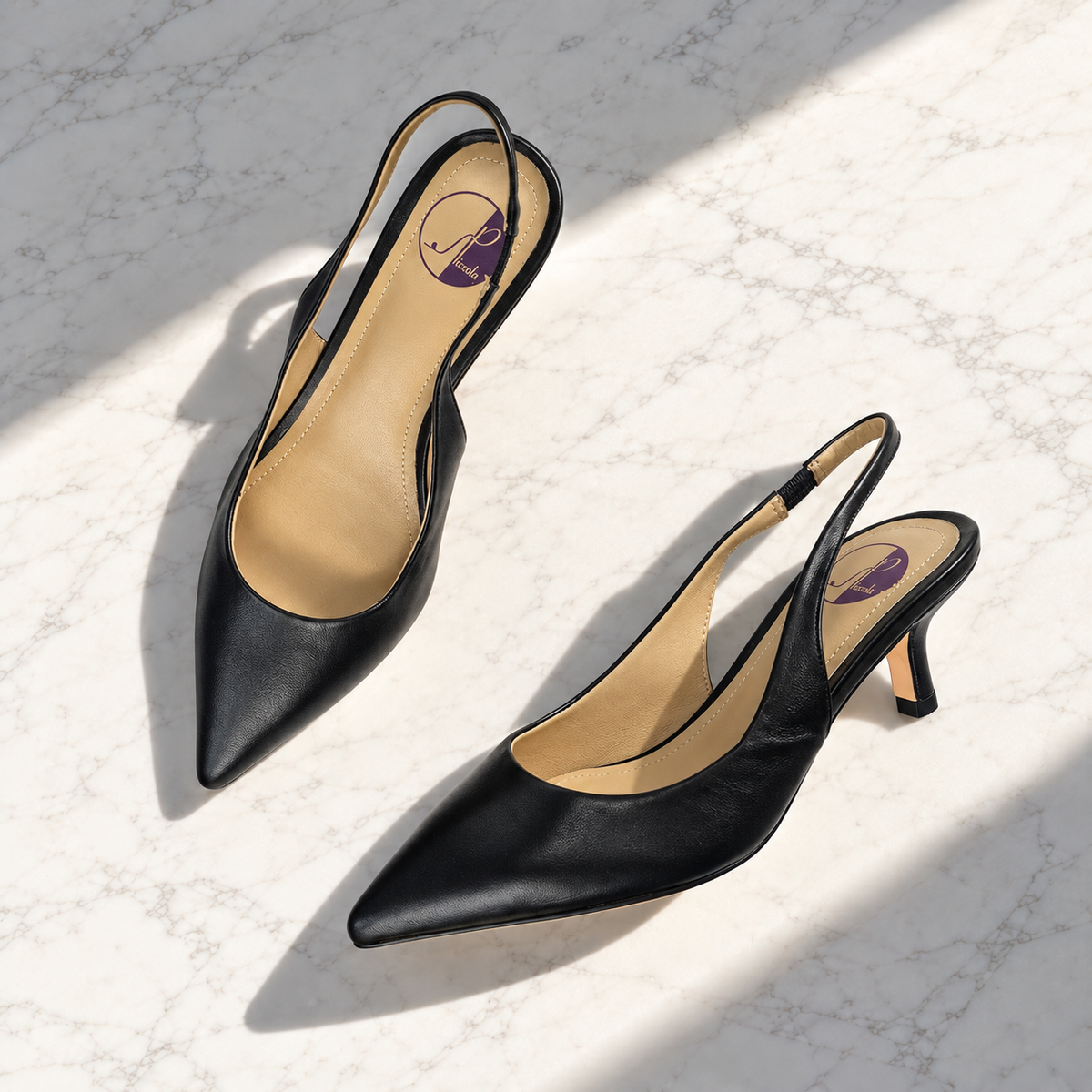 A pair of black leather pointed-toe slingback pumps with kitten heels, displayed on a light marble surface with diagonal shadows.