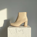 A beige leather ankle boot with a chunky block heel and a small pull tab at the back, shown from a side profile on a white studio background.