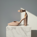 A single nude beige slingback high-heeled shoe with an ankle strap and block heel, displayed on a white marble pedestal against a gray background with a strong diagonal shadow.