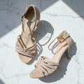 A pair of beige suede-like strappy high-heeled sandals with ankle straps, arranged on a white marble surface with sharp sunlight shadows.