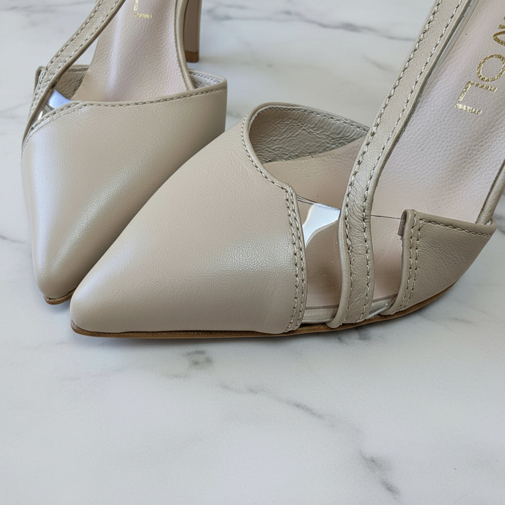Close-up of ORA nude leather heels showing transparent side panel and premium stitching detail on marble surface with foliage.