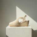 A single woven espadrille wedge sandal in a light beige tone, featuring elastic ankle straps, displayed on a white marble pedestal against a neutral gray wall with dramatic diagonal sunlight.