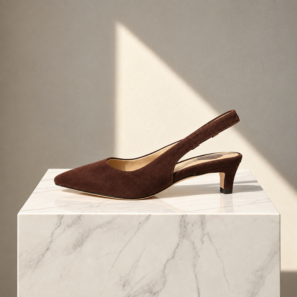 A single dark brown suede slingback pump with a pointed toe and low block heel, displayed on a white marble pedestal against a neutral beige wall with soft diagonal sunlight.