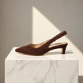 A single dark brown suede slingback pump with a pointed toe and low block heel, displayed on a white marble pedestal against a neutral beige wall with soft diagonal sunlight.