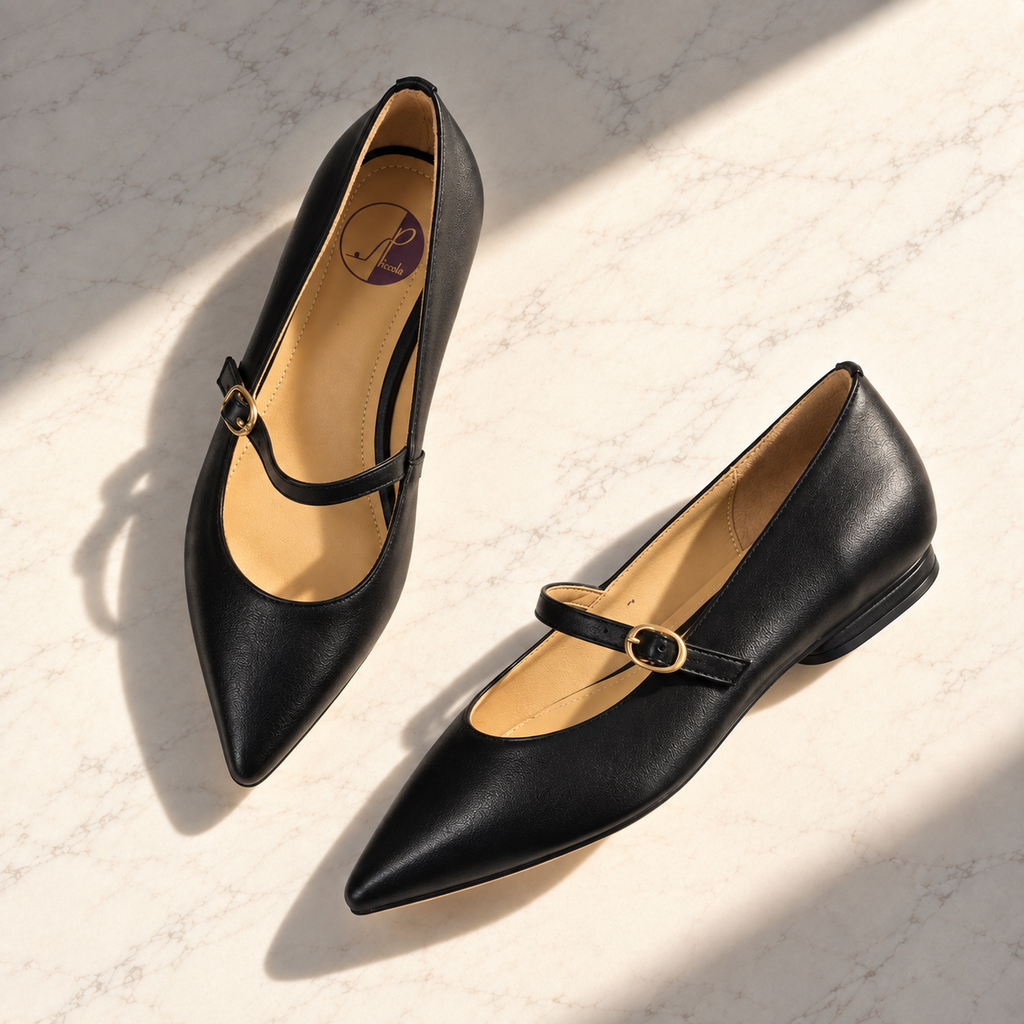 A pair of black leather pointed-toe Mary Jane flats with low block heels and gold-tone buckle straps, displayed on a light marble surface with diagonal shadows.
