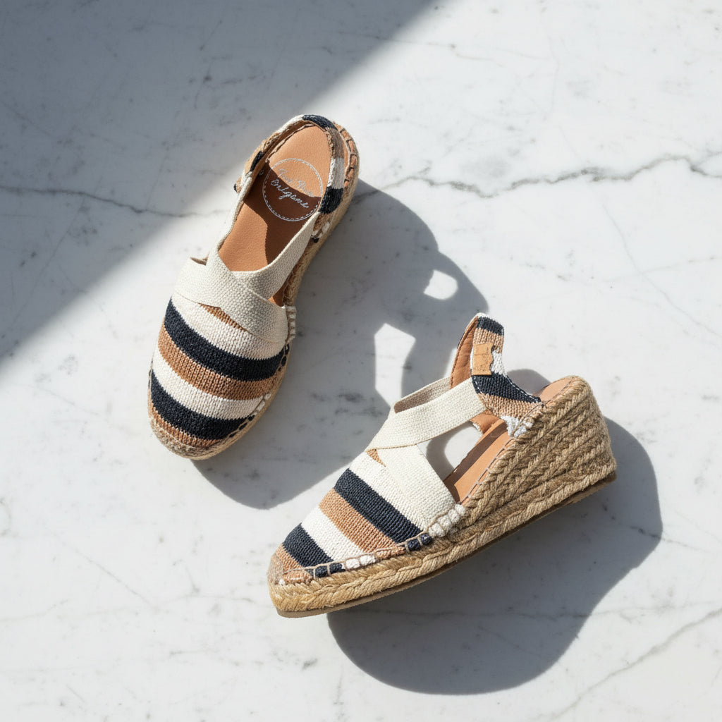 A pair of wedge espadrille sandals with woven jute wedge heels and striped uppers in cream, tan, and navy, photographed on a light marble surface with strong sunlight and shadows.