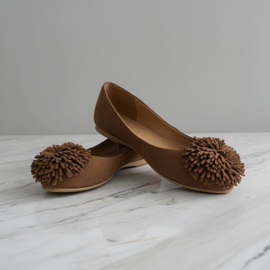 Brown flat shoes with pom-pom details on a light gray background