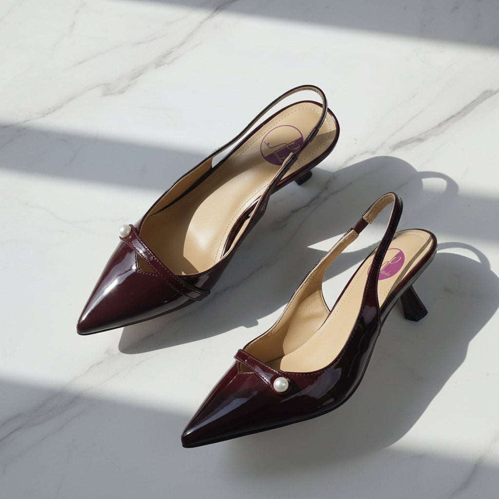 Pair of burgundy patent leather pointed-toe slingback heels with pearl embellishment laid flat on white marble surface with natural sunlight shadows.