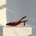 Single burgundy patent leather pointed-toe slingback kitten heel with pearl detail displayed in profile on white marble pedestal, light grey studio backdrop with soft sunbeam.