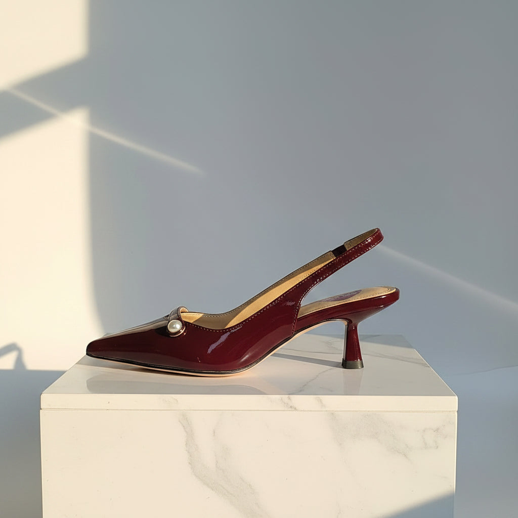 Single burgundy patent leather pointed-toe slingback kitten heel with pearl detail displayed in profile on white marble pedestal, light grey studio backdrop with soft sunbeam.