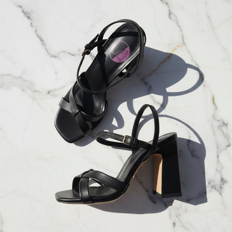Black leather strappy sandals set on a chunky block heel and presented on a marble background. 