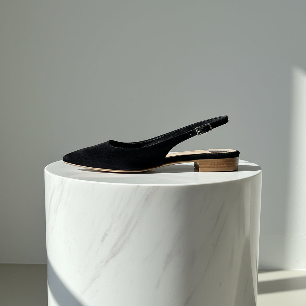 Black suede pointed toe slingback ballerina flats set on a marble stand.