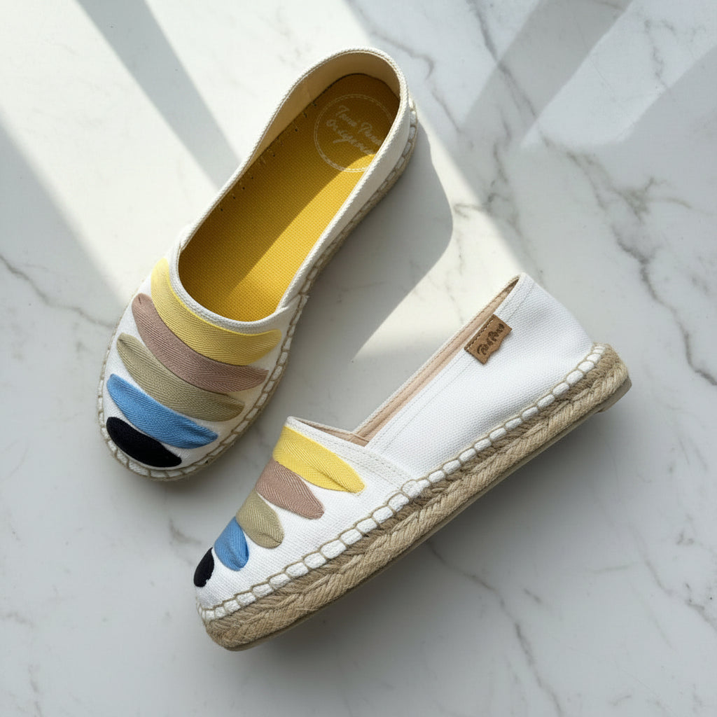 A pair of white espadrille slip-on shoes with colorful painted accents in yellow, beige, blue, and black, displayed on a white marble surface.