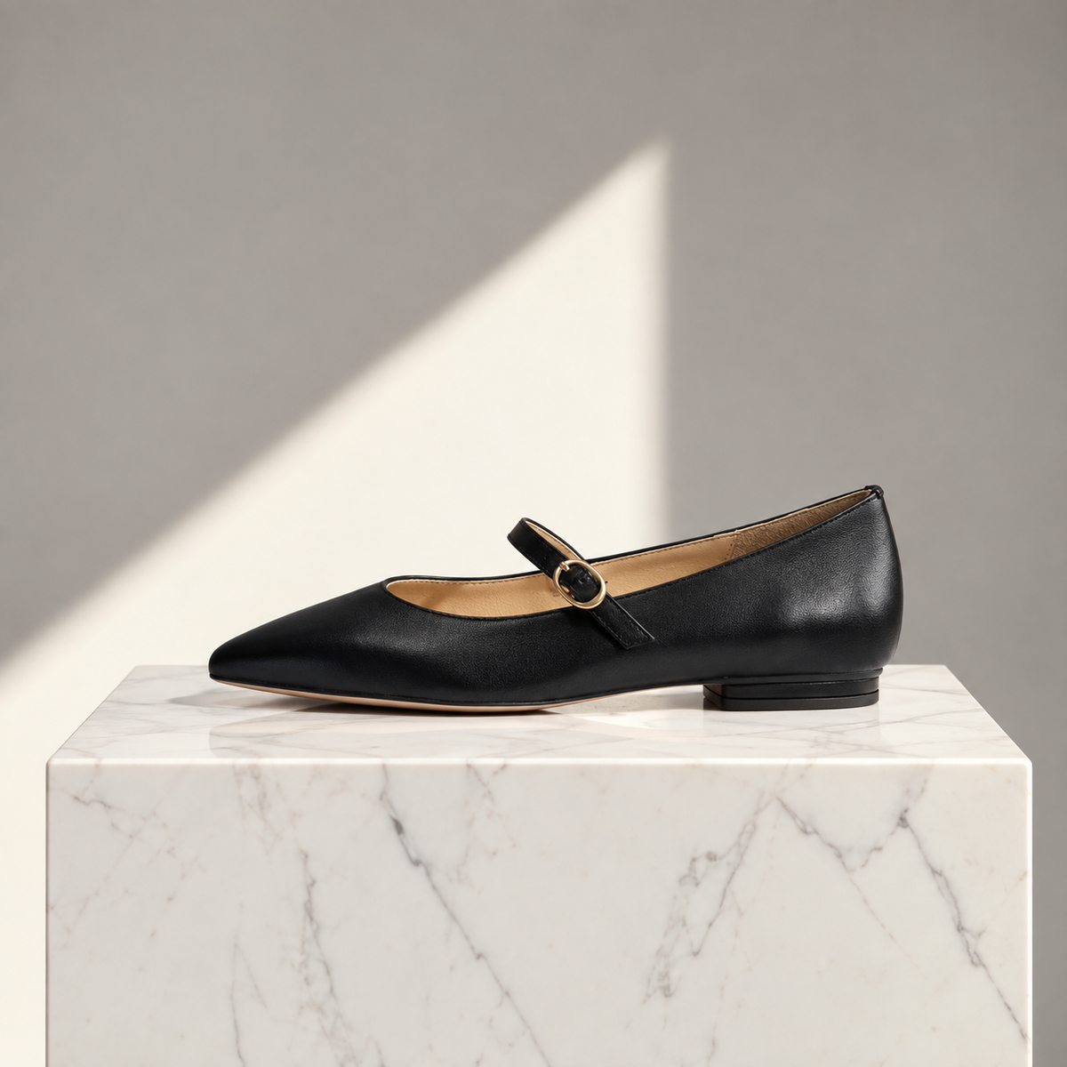 A single black leather Mary Jane flat shoe with a pointed toe and a gold-tone buckle, displayed on a white marble pedestal against a gray background with a diagonal light shadow.