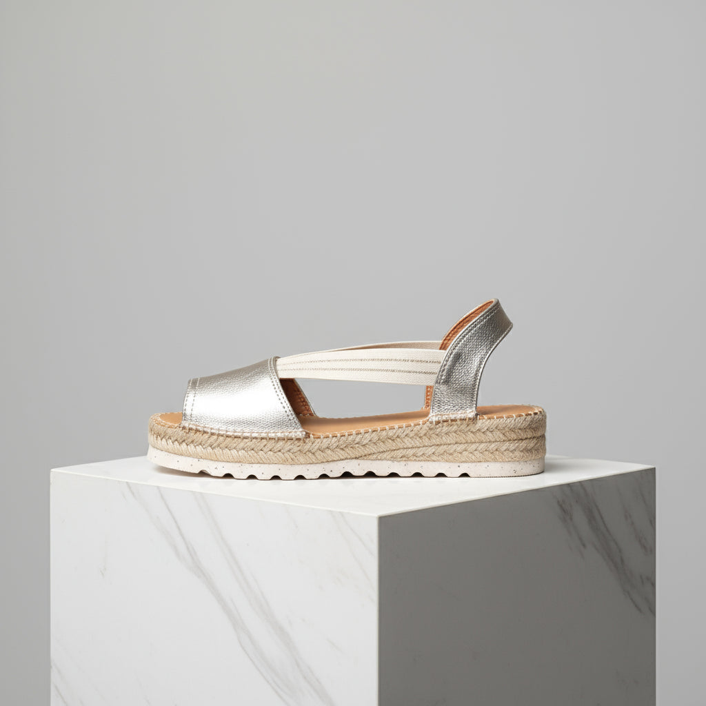 Gold sandal with espadrille sole on a white pedestal against a gray background