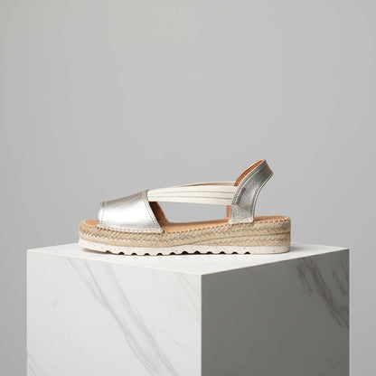 Gold sandal with espadrille sole on a white pedestal against a gray background