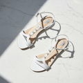 A pair of white leather flat sandals with thin straps, decorative pearl accents, and low block heels, displayed on a white marble surface in bright sunlight with sharp shadows.
