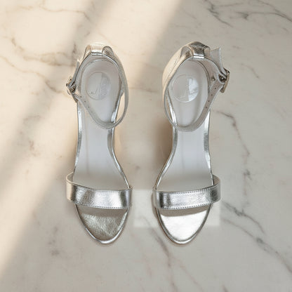 Front view of pair of silver high heel sandals with ankle straps on marble.