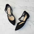 Black pointed-toe shoes with ankle straps on a white mat.