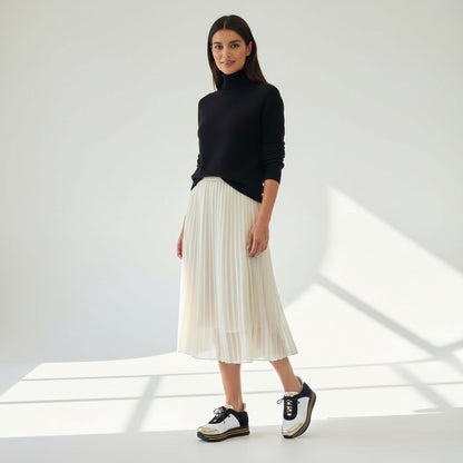 Woman wearing a black turtleneck, white pleated skirt, and black and white sneakers in a bright room.