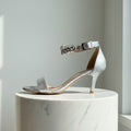 Silver high-heeled sandal with embellishments on a white marble pedestal against a light gray background