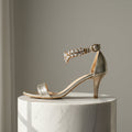 Gold high-heeled sandal with embellishments on a marble pedestal against a gray background