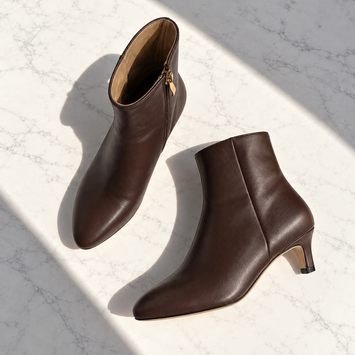 A pair of dark brown leather ankle boots with pointed toes and kitten heels, displayed on a light marble surface with dramatic diagonal shadows.