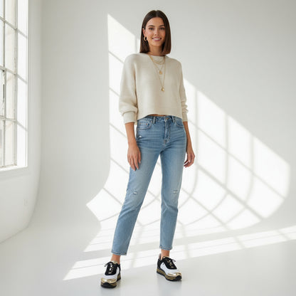 Woman wearing a white sweater and blue jeans standing in a bright room with sunlight filtering through.