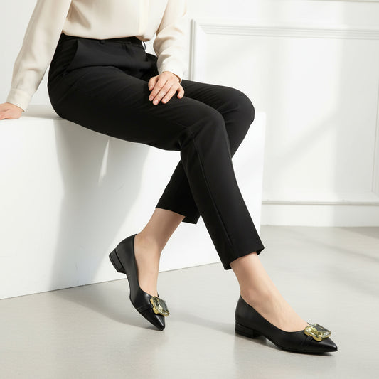 Person wearing black pants and black pointed toe ballerina shoes with gold accents sitting on a white surface.