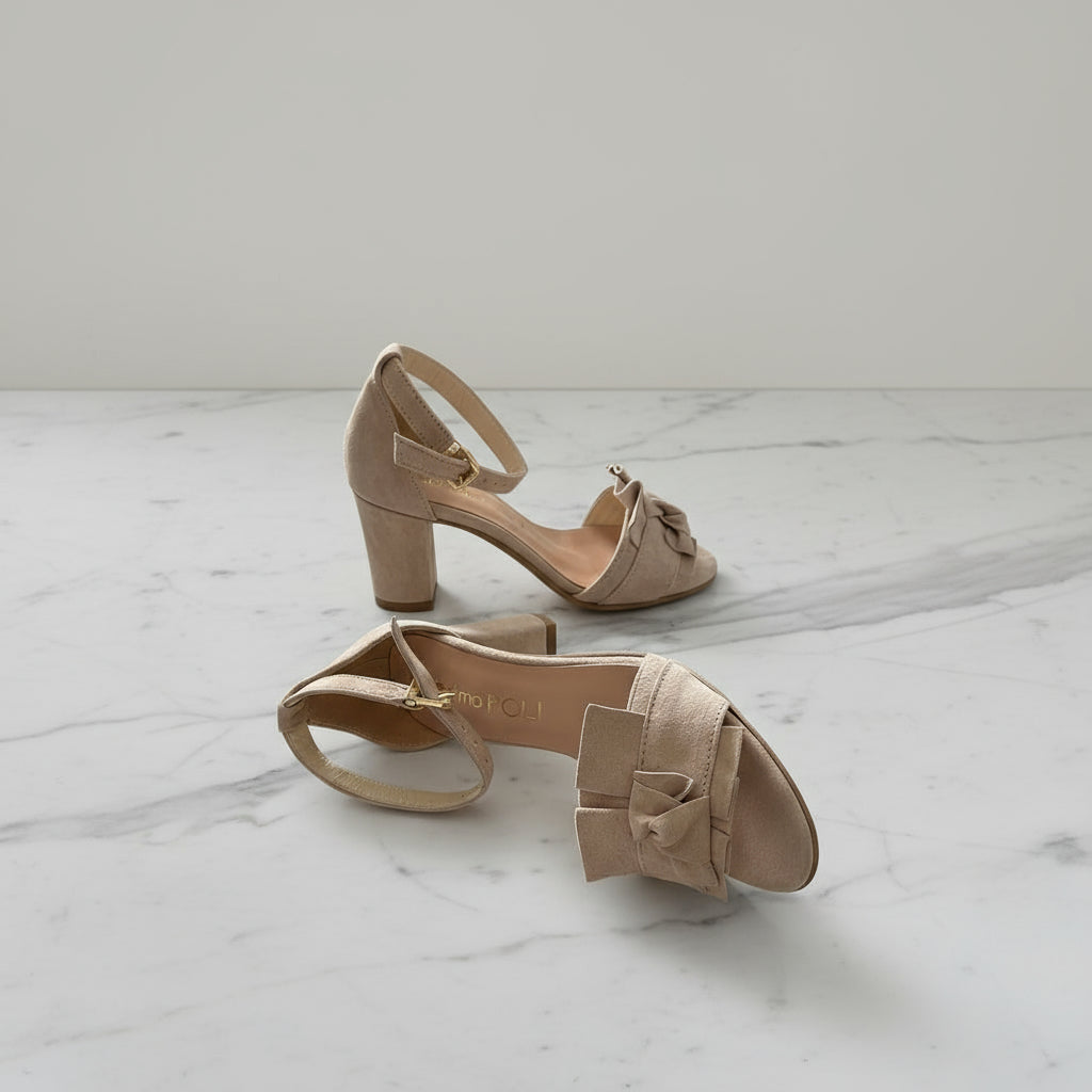 Pair of beige high-heeled sandals with bow details on a marble surface.